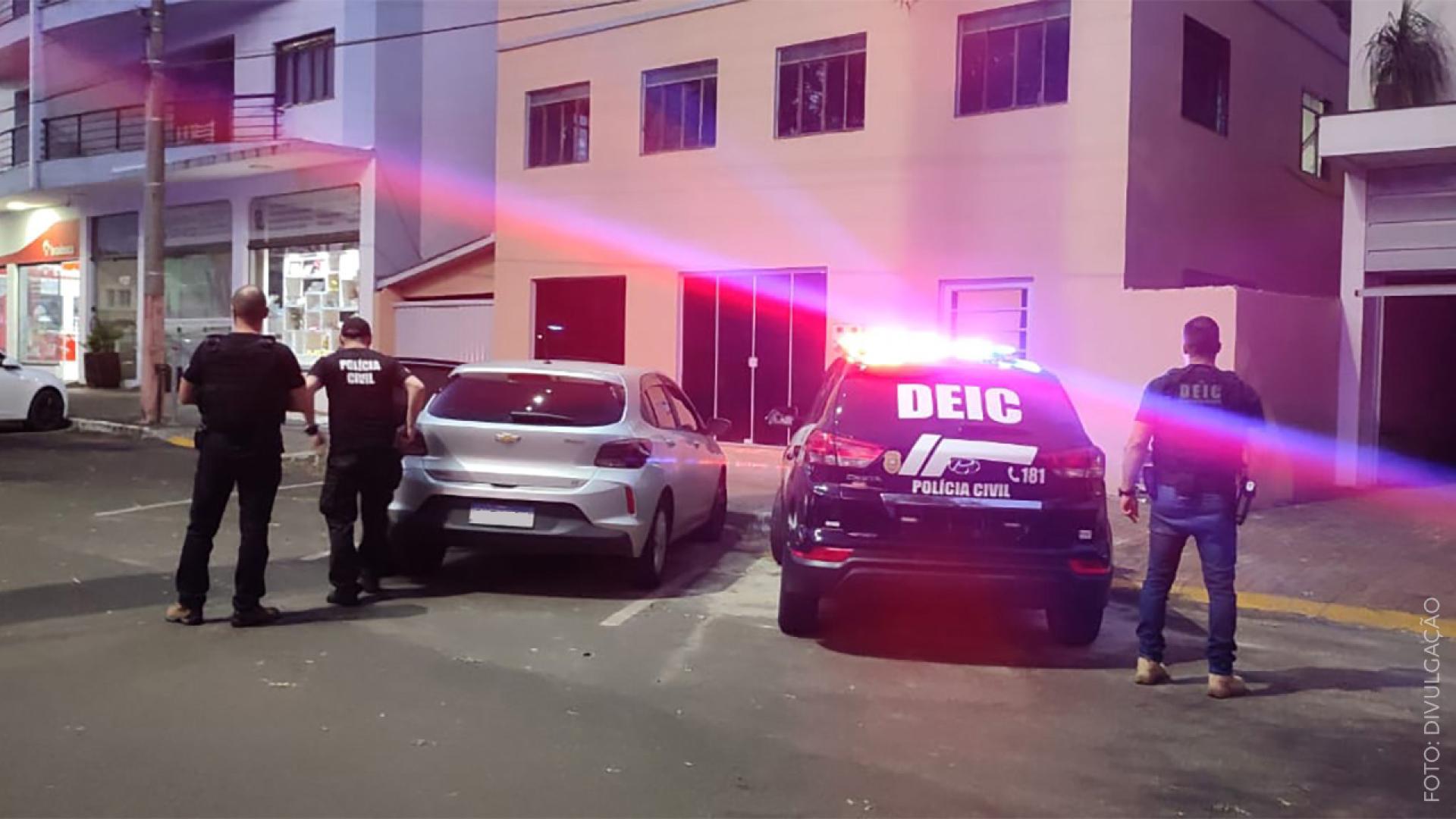 Imagem mostra veículo da polícia civil com luzes de emergência ligadas parado em frente a um prédio. Há três policiais de costas na imagem. 