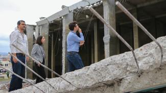 Imagem mostra três pessoas observando área destruída. São dois homens e uma mulher. Em primeiro plano, aparecem escombros com vigas de ferro à mostra.
