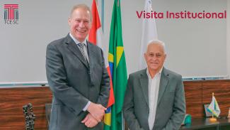 Foto de dois homens brancos, ambos de ternos cinzas. Ao fundo, as bandeiras do Brasil e de Santa Catarina. No alto, à direita, está escrito “Visita Institucional”.