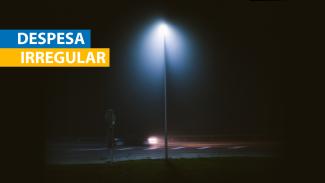 Banner horizontal com a imagem de uma rua, à noite, iluminada por um poste de luz, ao centro. Na lateral esquerda superior, o texto “Despesa irregular”, em fonte branca e em retângulos azul e amarelo. 